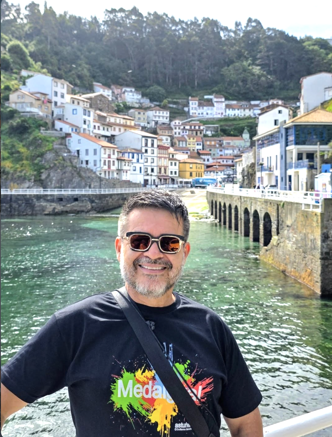 Hombre con gafas de sol sonriendo junto a un puerto con casas de colores en una ladera verde al fondo